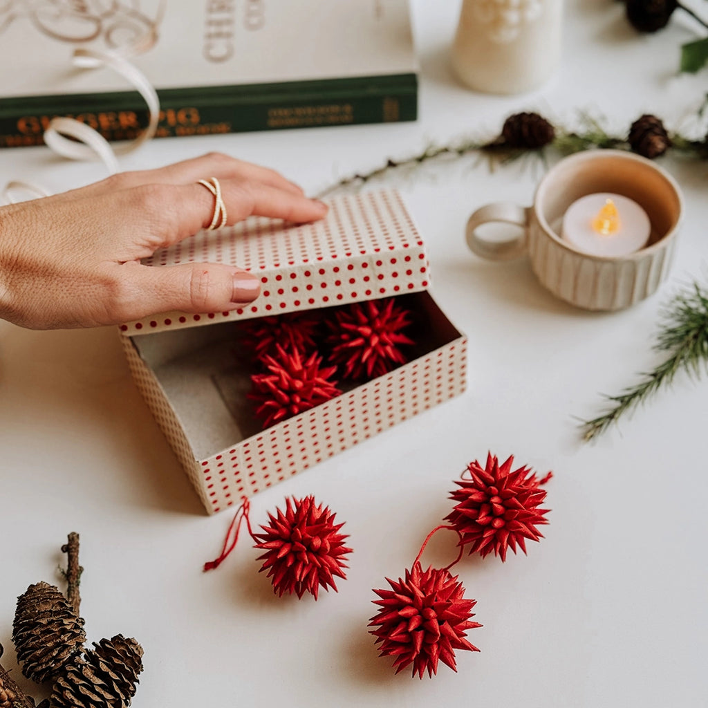 A hand is lifting the lid off of a box of red quilled paper Christmas decorations. a tea light, pine cones and a Christmas recipe book are in the background.