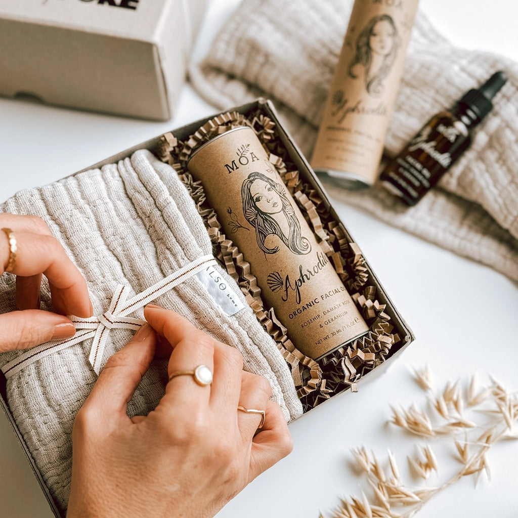 A woman's hands tying a ribbon around a wash cloth inside byFoke's Ingrid Gift Box, which is laying open on a table. The gift box also contains Aphrodite Organic Facial Oil by Magic Organic Apothecary.