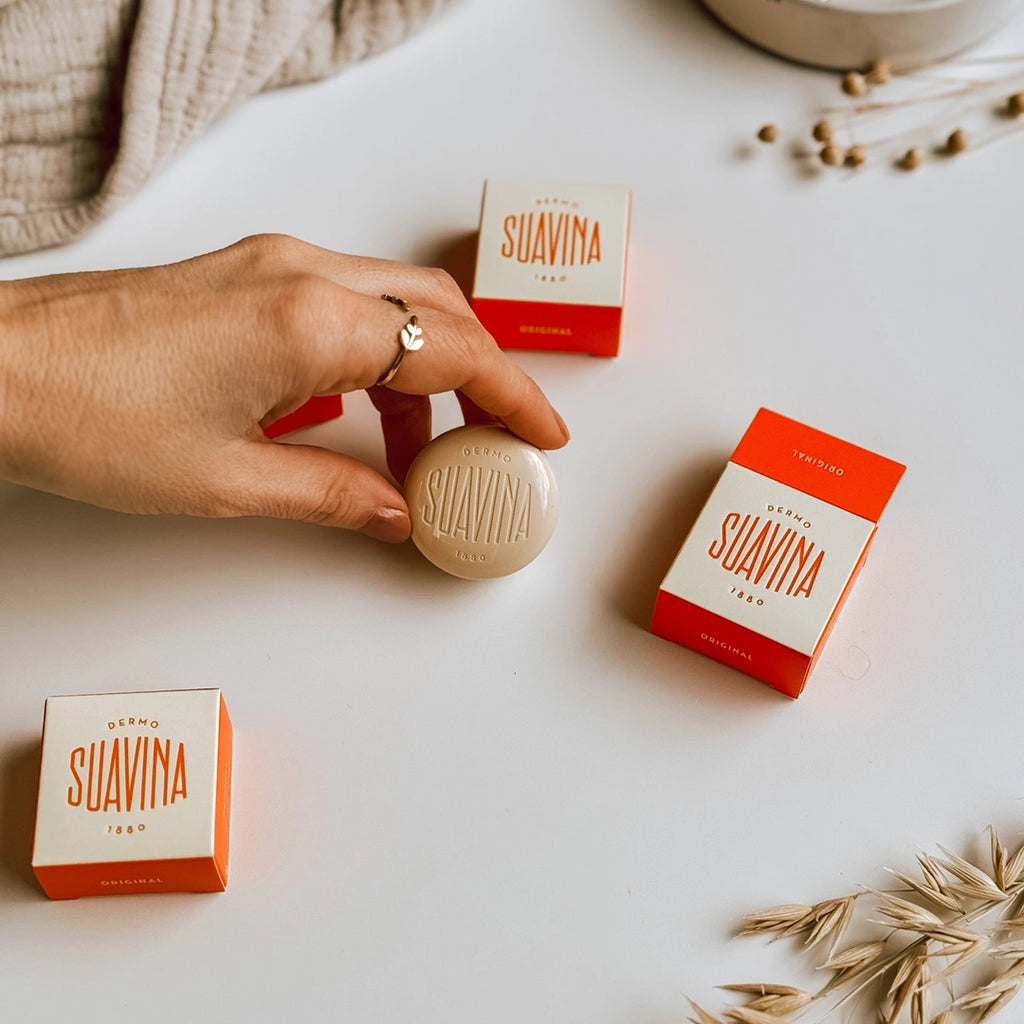 A lady's hand picking up the Dermo Suavina Lip Balm pot with some of the cream and orange boxes of lip balm in the background.