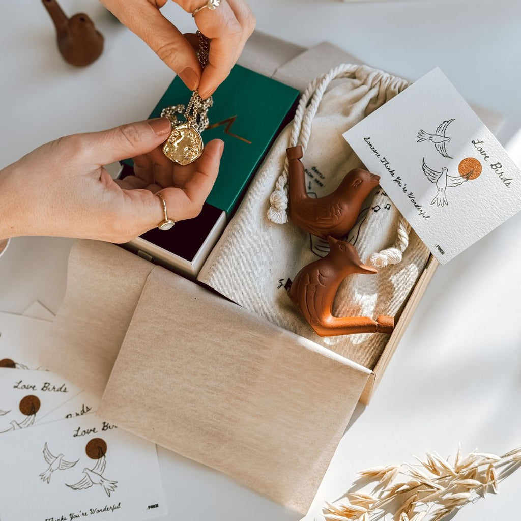 Hands holding up a gold necklace above a byFoke gift box that also contains a pair of ceramic birds and an illustrated love birds note card.