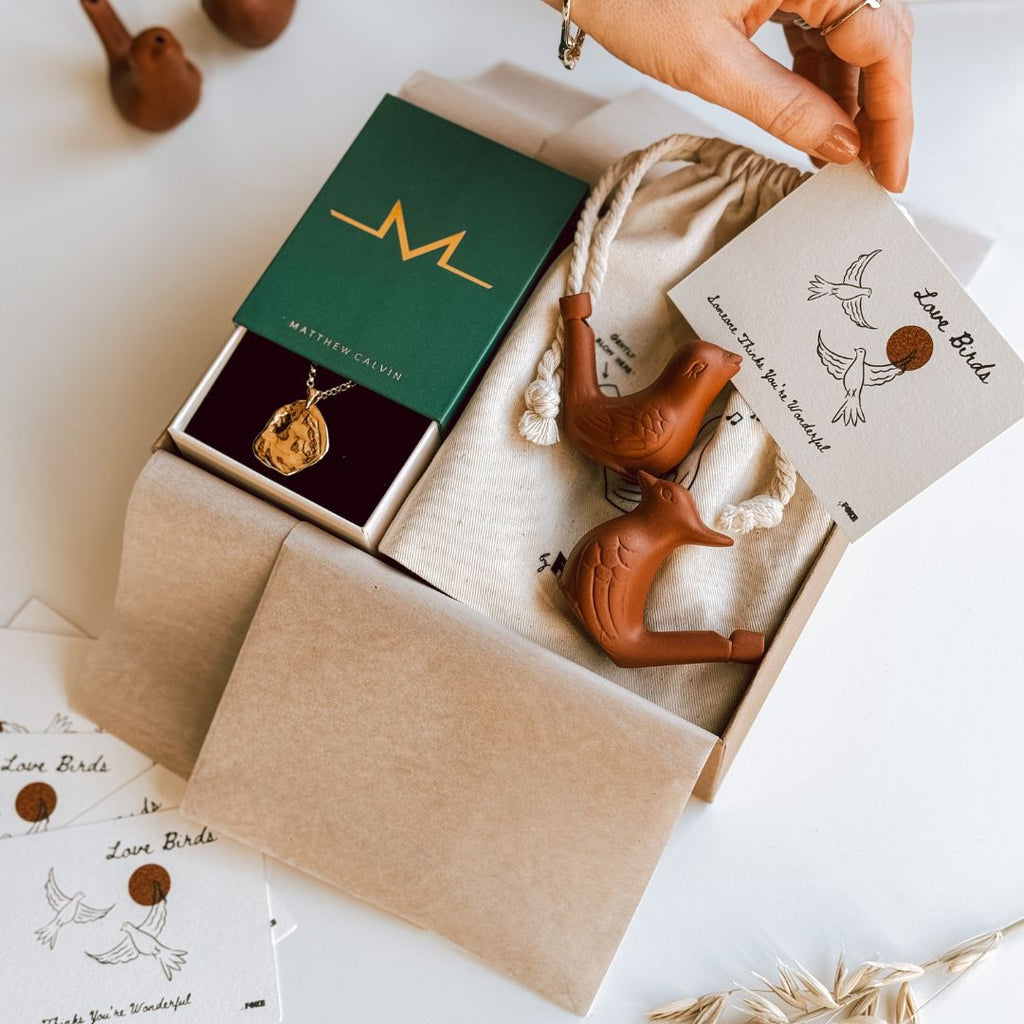 An illustrated love birds note card being placed into an open byFoke gift box that contains a gold necklace and a pair of ceramic bird whistles.