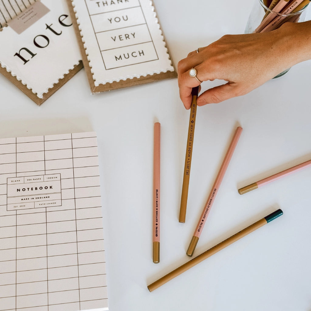 A hand lifting a Katie Leamon pencil up from a table with other pencils and notecards in the background.