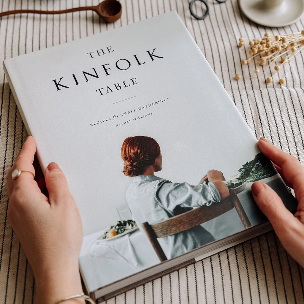 The Kinfolk Table Recipes for Small Gatherings Cook Book on a striped table cloth in a woman's hands. byFoke