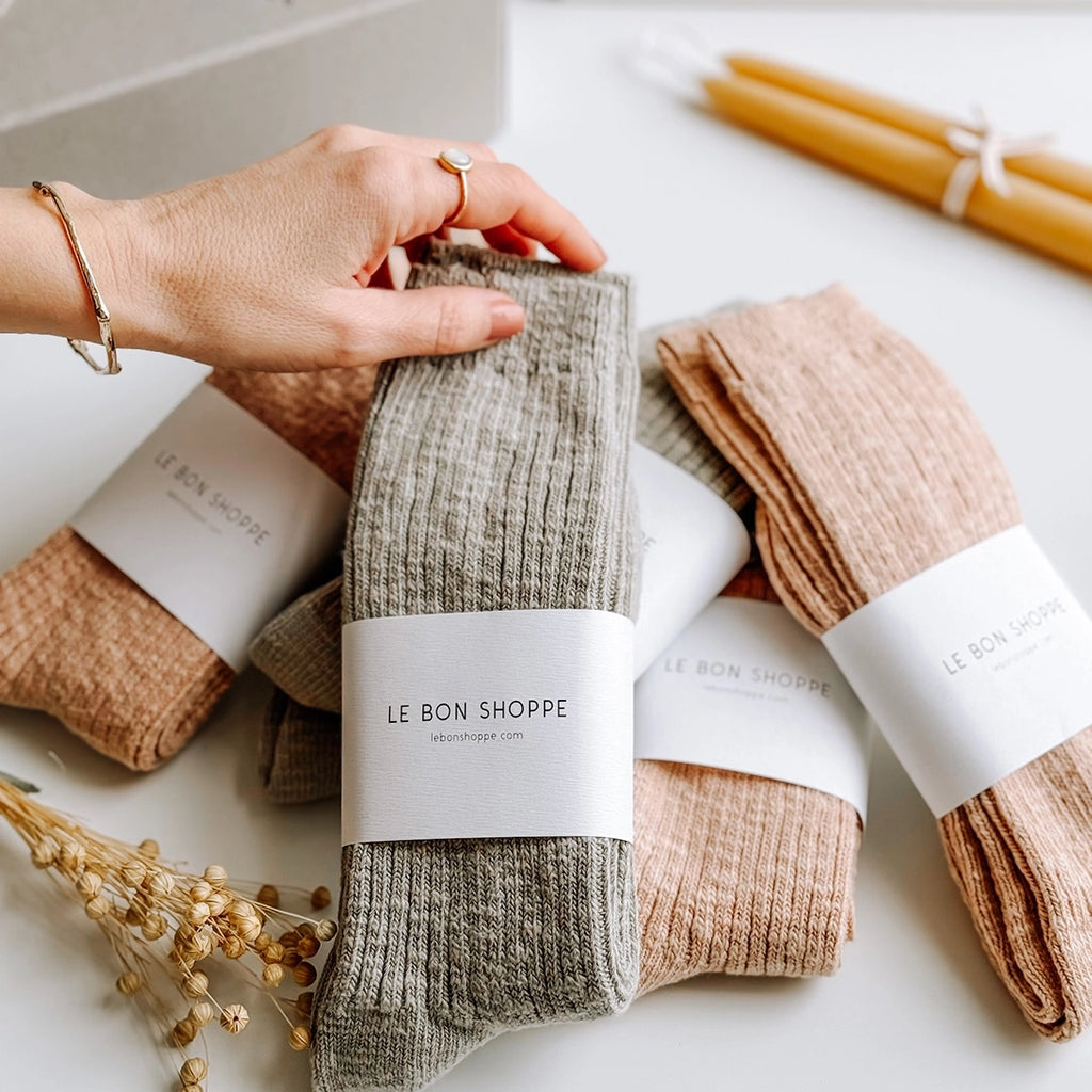 A pile of Le Bon Shoppe Cottage Socks in Smoked Sage and Peachy Keen. A woman's hand is placing one of the pairs of socks in the pile. byFoke.