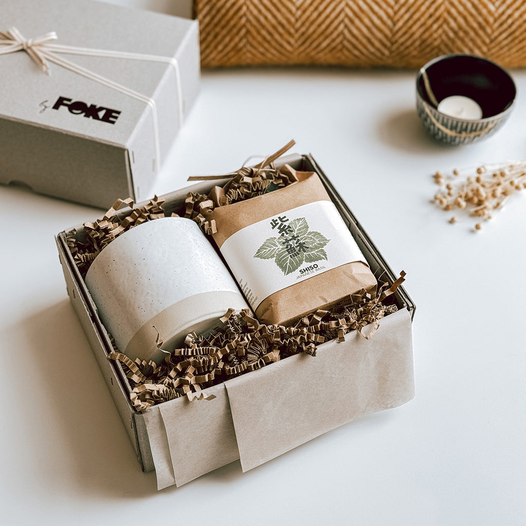 The Maiko gift box byFoke laying open on a table showing the contents, which are a Japanese Herb Growing Kit and a ceramic plant pot. The lid of the gift box is laying to one side wrapped in ribbon.