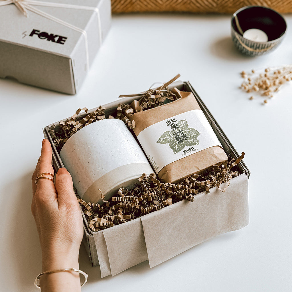 A woman's hand placing a Maiko gift box byFoke onto a table. The gift box is open showing the contents, which are a Japanese Herb Growing Kit and a ceramic plant pot.
