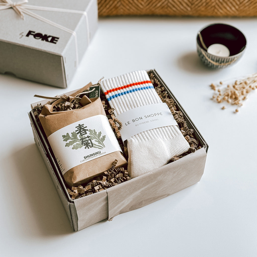 The Mei Gift Box byFoke, open on a table with a blanket and a candle in the background. The box contains a pair of Le Bon Shoppe Socks that are cream with a red and blue stripe and a cultivate and eat herb kit.