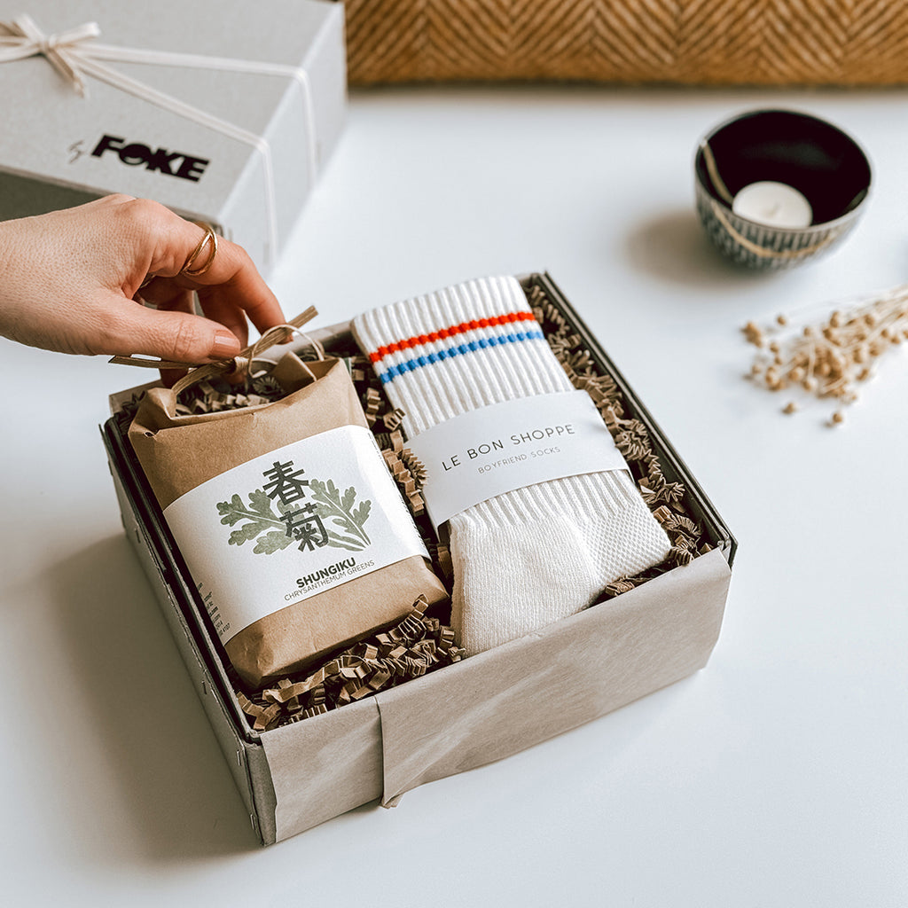 The Mei Gift Box byFoke, open on a table. The box contains a pair of Le Bon Shoppe Socks that are cream with a red and blue stripe and a cultivate and eat herb kit.