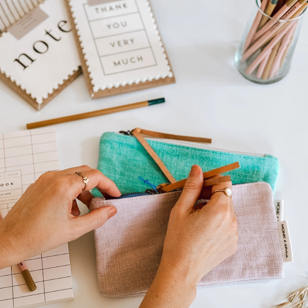 Hands opening the zip of a Monk & Anna pink linen pencil case which is on a table in the byFoke studio with a green pencil case and other pencils and notecards in the background.