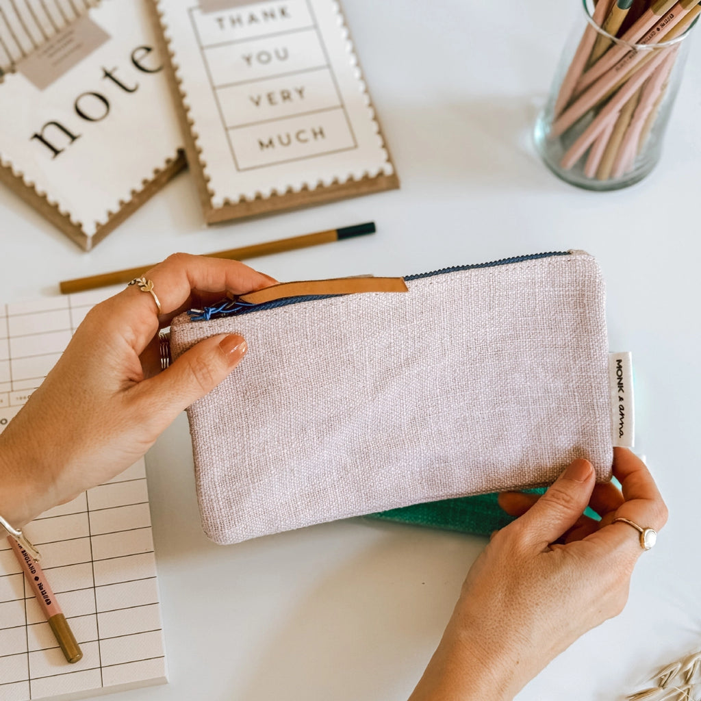 Hands holding a Monk & Anna pink linen pencil case in the background there are pencils and notecards.