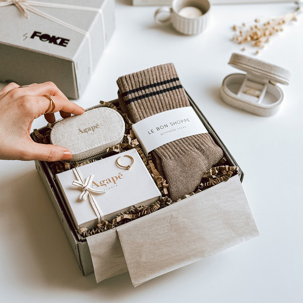 byFoke's Romy Luxury Gift Box laying open on a table with the lid in the background. Inside you can see an Agape gold ring, linen jewellery box and a pair of Le Bon Shoppe socks in Cocoa. A woman's hand is placing the jewellery box inside the gift box.