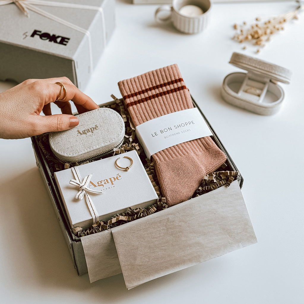 byFoke's Romy Luxury Gift Box laying open on a table with the lid in the background. Inside you can see an Agape gold ring, linen jewellery box and a pair of Le Bon Shoppe socks in Vintage Pink. A woman's hand is placing the jewellery box inside the gift box.