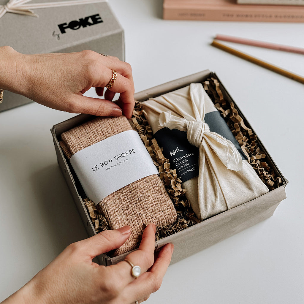 The Runa Gift Box byFoke, laying open on a table with a woman's hands arranging the socks inside the box. The Gift box contains a pair of Le Bon Shoppe Cottage Socks in Peachy Keen and a bar of Harth Cornish Sea Salt Chocolate.