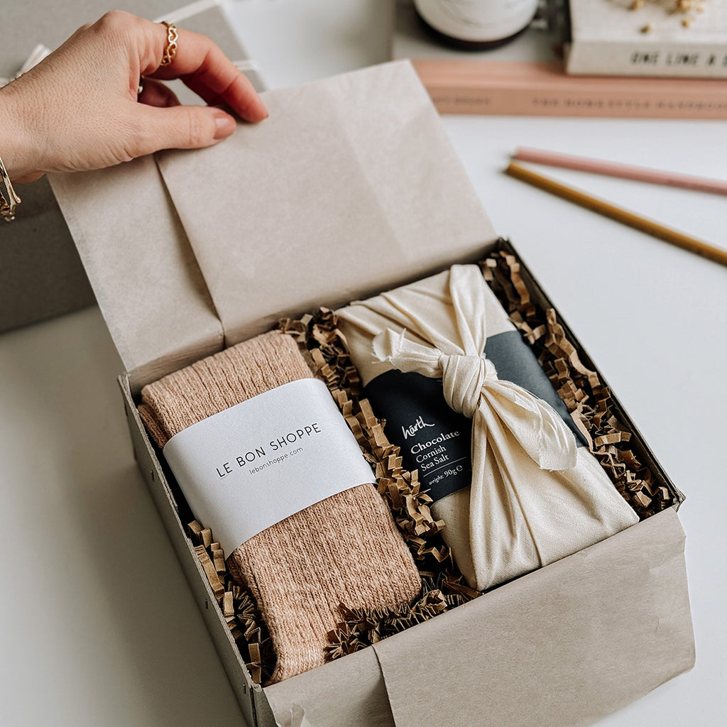 The Runa Gift Box byFoke, being opened by a woman's hand. The Gift box contains a pair of Le Bon Shoppe Cottage Socks in Peachy Keen and a bar of Harth Cornish Sea Salt Chocolate.