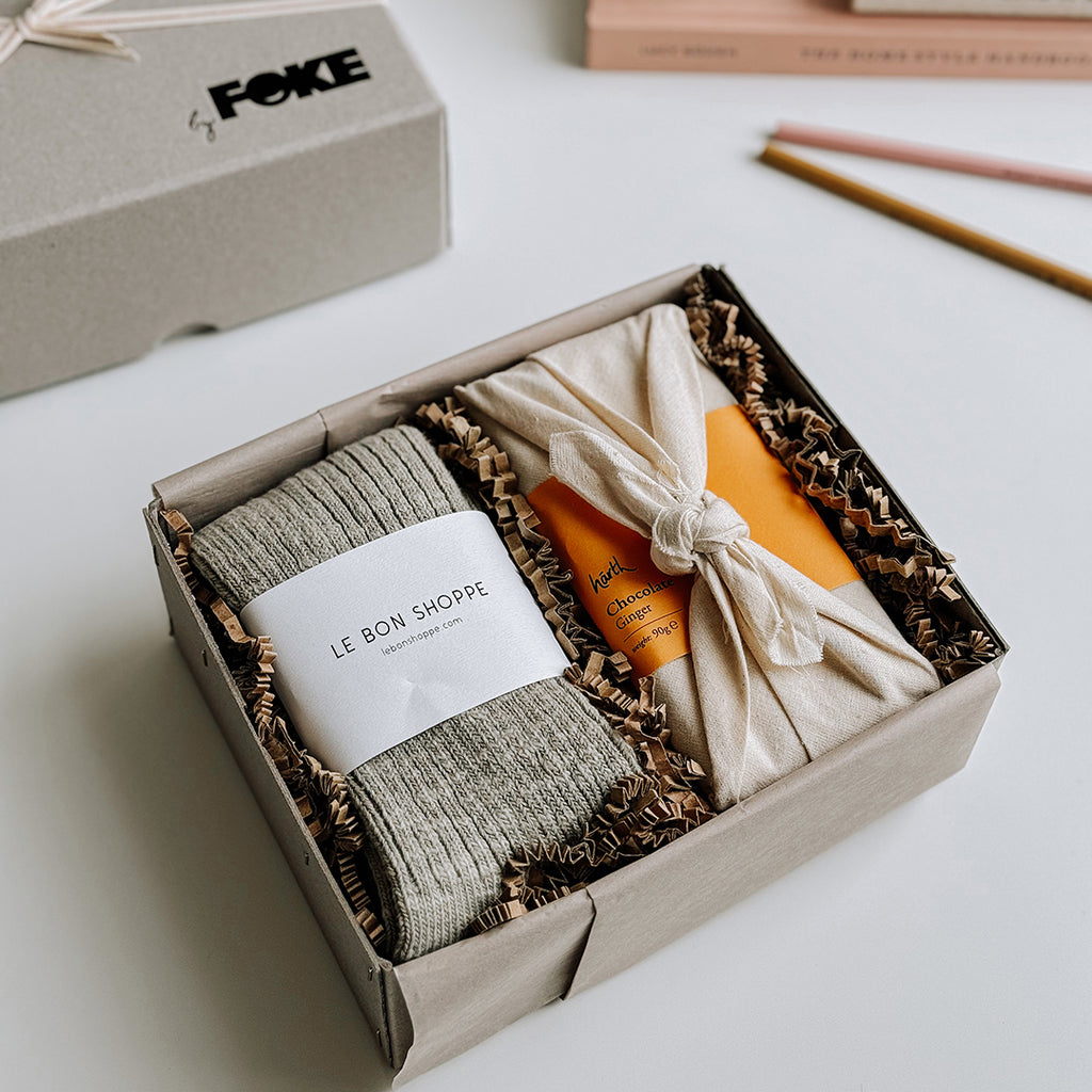 The Runa Gift Box byFoke, laying open on a table with the lid to one side. The Gift box contains a pair of Le Bon Shoppe Cottage Socks in Smoked Sage and a bar of Harth Ginger Chocolate.