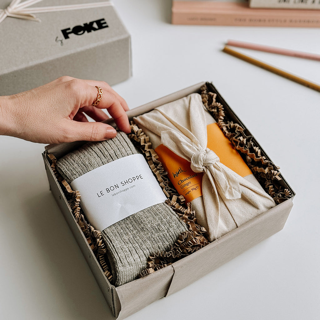 The Runa Gift Box byFoke, laying open on a table. The Gift box contains a pair of Le Bon Shoppe Cottage Socks in Smoked Sage and a bar of Harth Ginger Chocolate.
