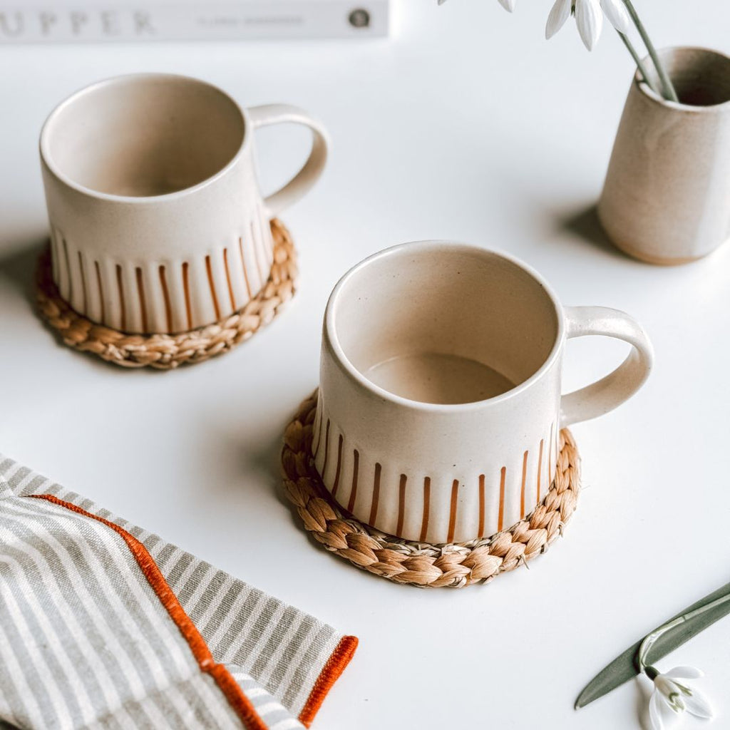 A pair of striped stoneware mugs that are cream with a ginger stripe on woven coasters on a table, byFoke.