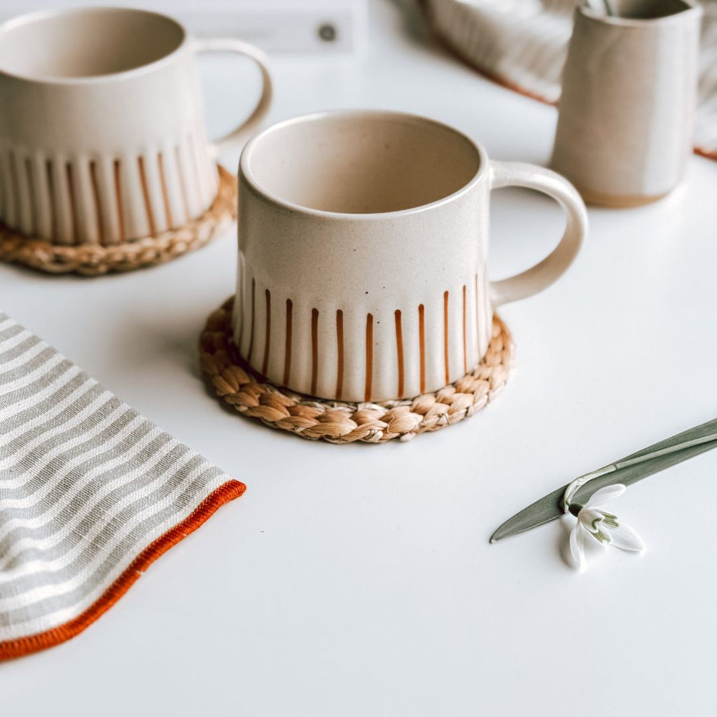 A close up of a striped stoneware mug which is cream with a ginger stripe on a woven coaster on a table, byFoke.