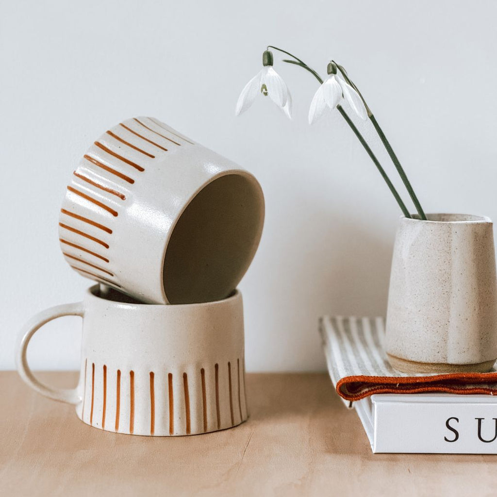 A stacked pair of striped stoneware mugs on a shelf with a book and some snowdrops in a vase, byFoke.