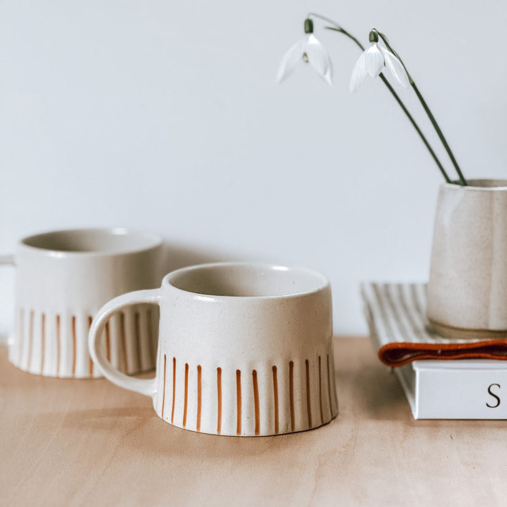 A pair of striped stoneware mugs on a shelf with a book and some snowdrops in a vase, byFoke.