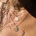 A close up photo of a woman's neck, with three necklaces from Agape Studio layered together. One of the necklaces is the Alba necklace, by Agape Studio which is a round gold pendent.