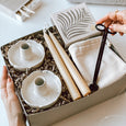 A woman's hands holding an open byFoke Cora gift box containing a set of two stoneware flower candle holders, a pair of beeswax taper candles, luxury letterpress matches by Archivist. The woman is lifting a pair of candle wick trimmers in her right hand..