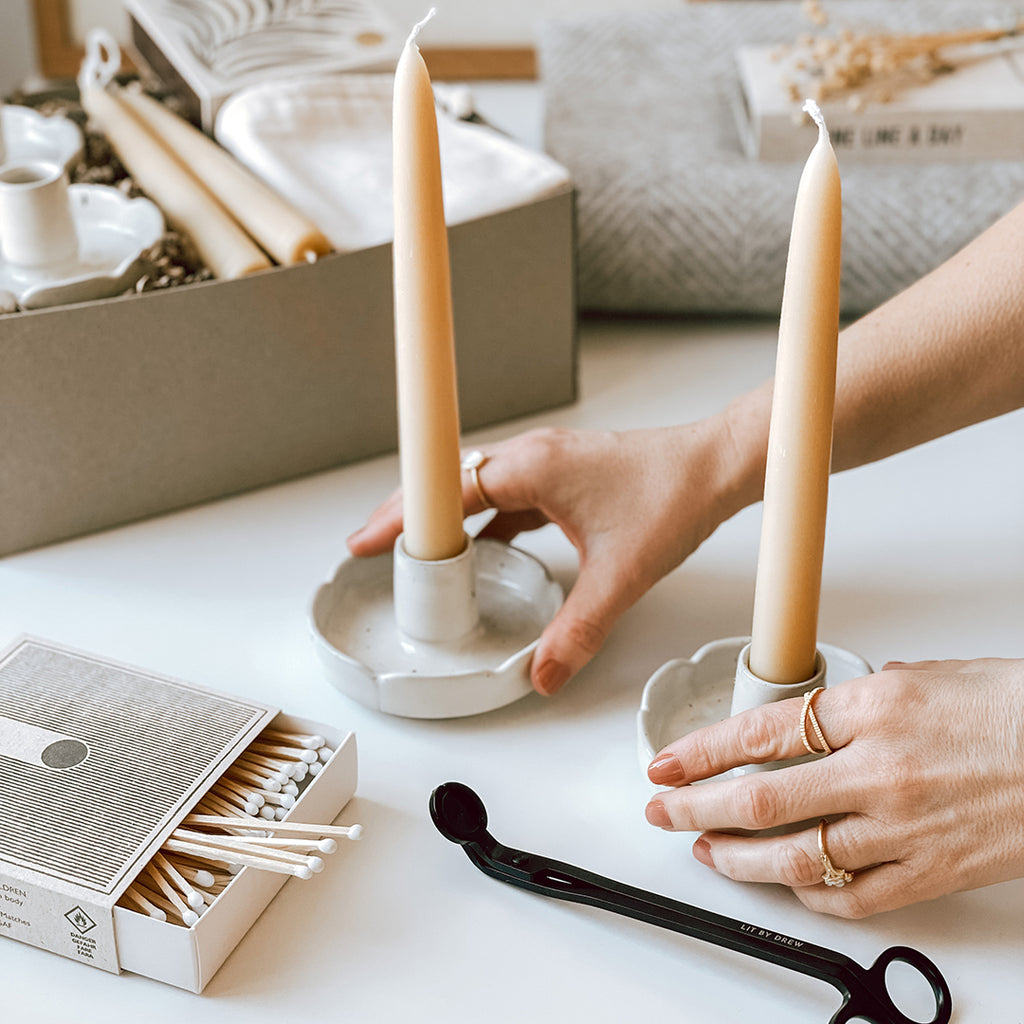 A woman's hands a placing a pair of stoneware flower candle holders containing beeswax candles onto a table with an open box of letterpress matches and a pair of candle wick trimmers in the foreground. In the background is an open byFoke Cora gift box and a byFoke blanket.