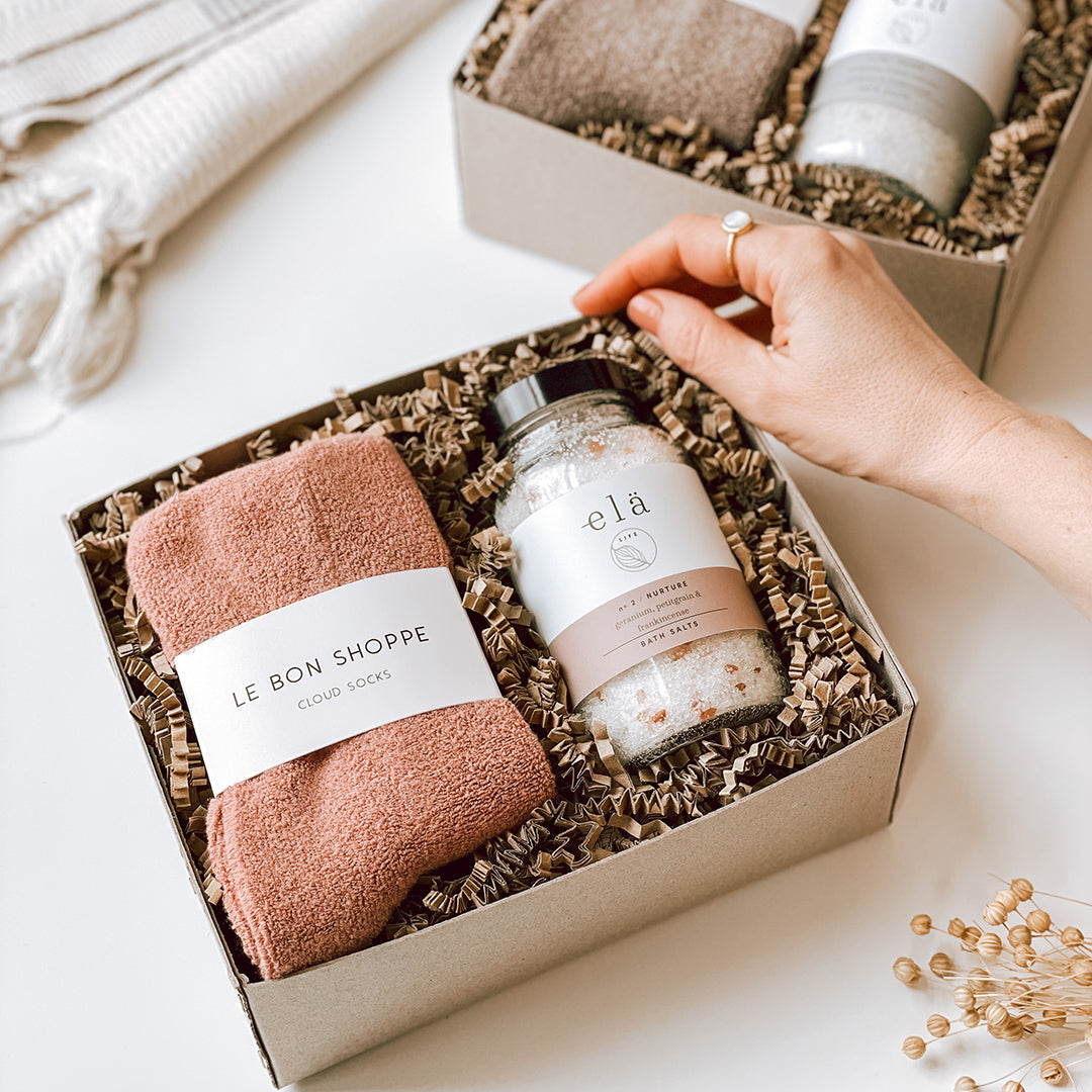 The byFoke Erin Gift Box in the Mulberry colour way, showing the open box held by a woman's hand, containing mulberry cloud socks by Le Bon Shoppe and Ela Life Bath Salts in Nurture.