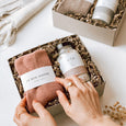 A woman's hands arranging the contents of the byFoke Erin Gift Box. The box contains mulberry cloud socks by Le Bon Shoppe and Ela Life Bath Salts in Nurture.