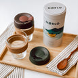 A bamboo tray laid with two glass Brew Cork KeepCups one with the lid off, an illustrated tin of Norlo coffee and a hand carved wooden spoon resting on a striped napkin.