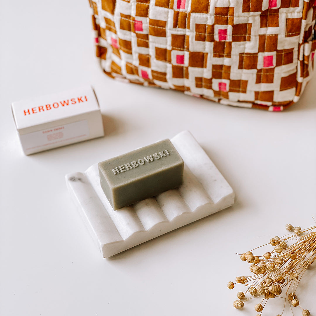 A Herbowski soap bar sitting on a carved marble soap dish with a block-rinted wash bag with a pink and rust print in the background.