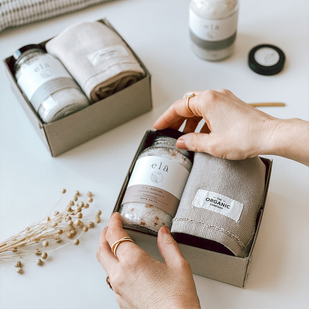Two byFoke Maeve Gift Boxes containing Ela Life Essential Oil Bath Salts and a calm wash cloth by The Organic Company. A woman's hands are arranging the contents of the open gift boxes.