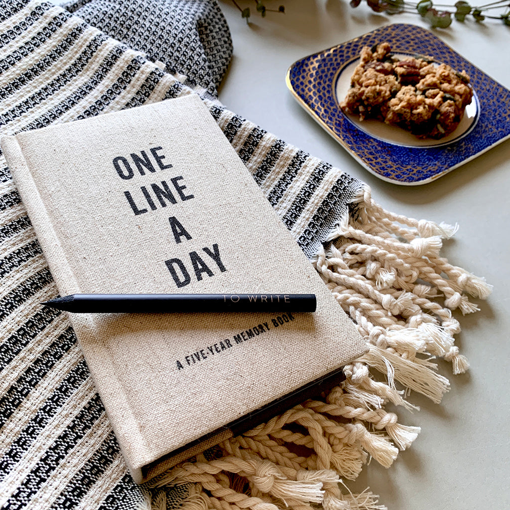 A linen covered One Line A Day 5 Year Memory Book laying on a black and white striped throw, with a small black pencil lying on top. In the background there is a homemade flapjack on a blue plate.