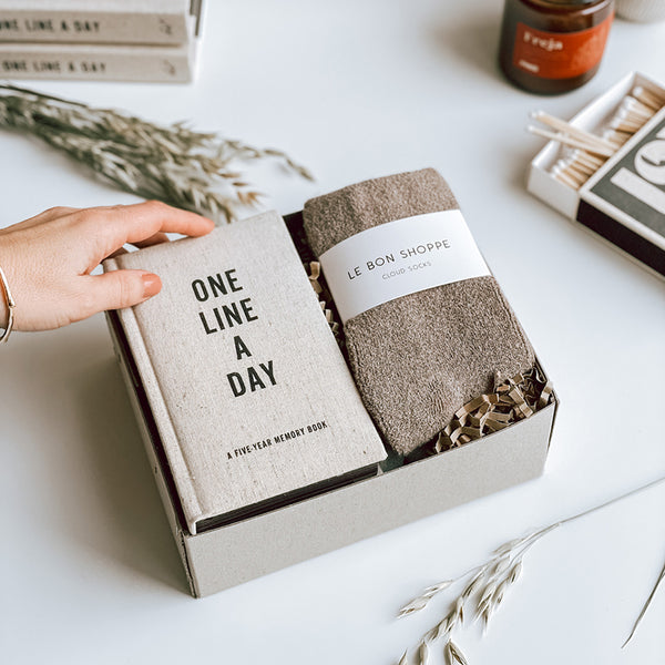 A byFoke Ottilie gift box containing a One Line a Day Journal and a pair of Le Bon Shoppe Cloud socks in Frappe. A ladies hand is lifting the corner of the journal our of the box.