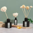 A photo of a collection of three reed diffusers by Ela Life.  Featured are the chrysanthemum diffuser, the pom pom diffuser and the flower travel diffuser. All of the diffusers are made of essential oils in glass bottles with handmade wooden flowers acting as the reeds. The diffusers are sitting on small slabs of stone with a teal background.