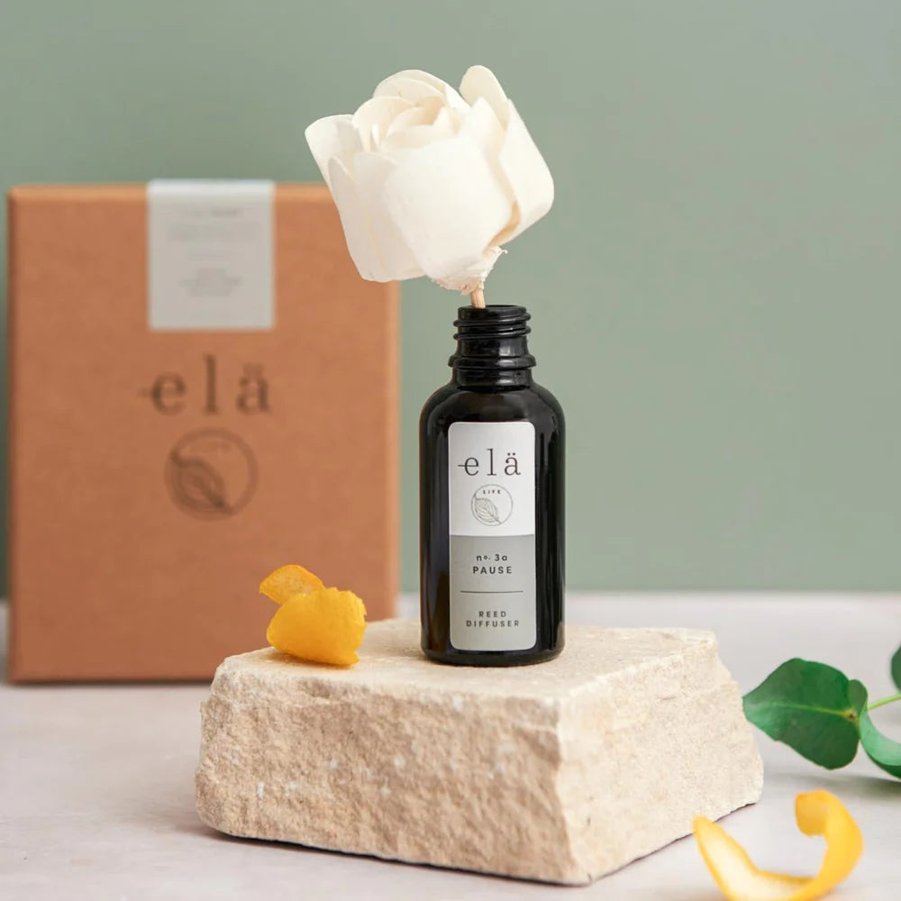 A small reed diffuser, with a handmade wooden flower to act as the reeds. The room diffuser is made up of essential oils in a glass apothecary bottle. The brand of the diffuser is Ela Life and the scent is Pause no. 3a. The diffuser is sitting on a small slab of stone with the sustainable gift box in the background.