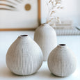 A close up of a collection of three beautiful byFoke ceramic bud vases all of slightly different sizes. The taller of the vases containsing a sprig of dried oats. Each vase is an ecru colour with a black dotty pattern printed onto it. The vases sit on a table with a framed print and a couple of books in the background.